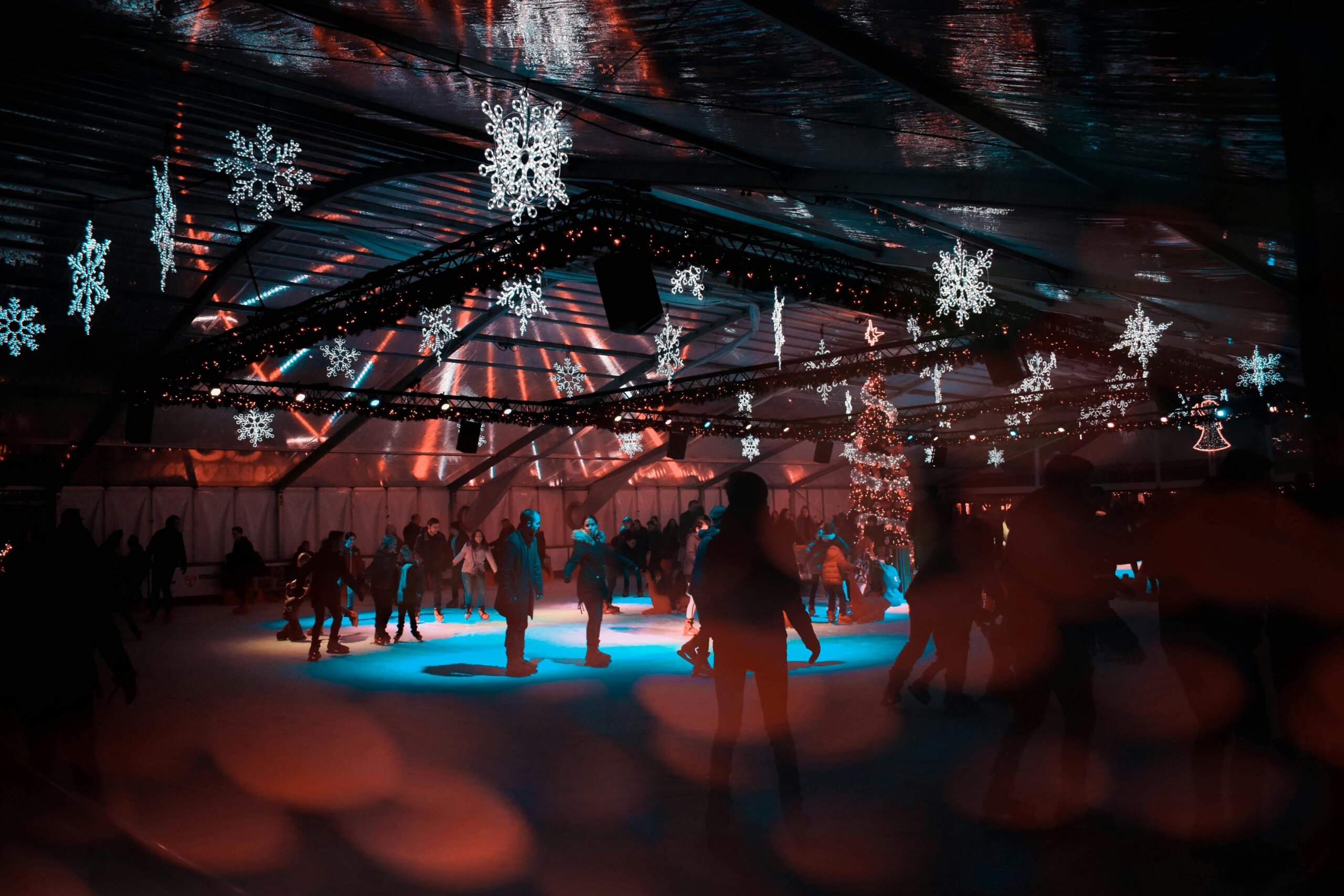 Indoor ice skating rink with people skating under festive snowflake lights, creating a joyful and vibrant atmosphere.