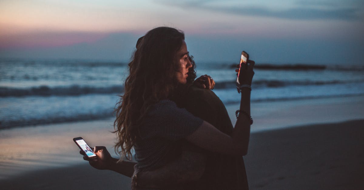 Couple embracing on a beach at sunset, using mobile phones, symbolizing modern love.