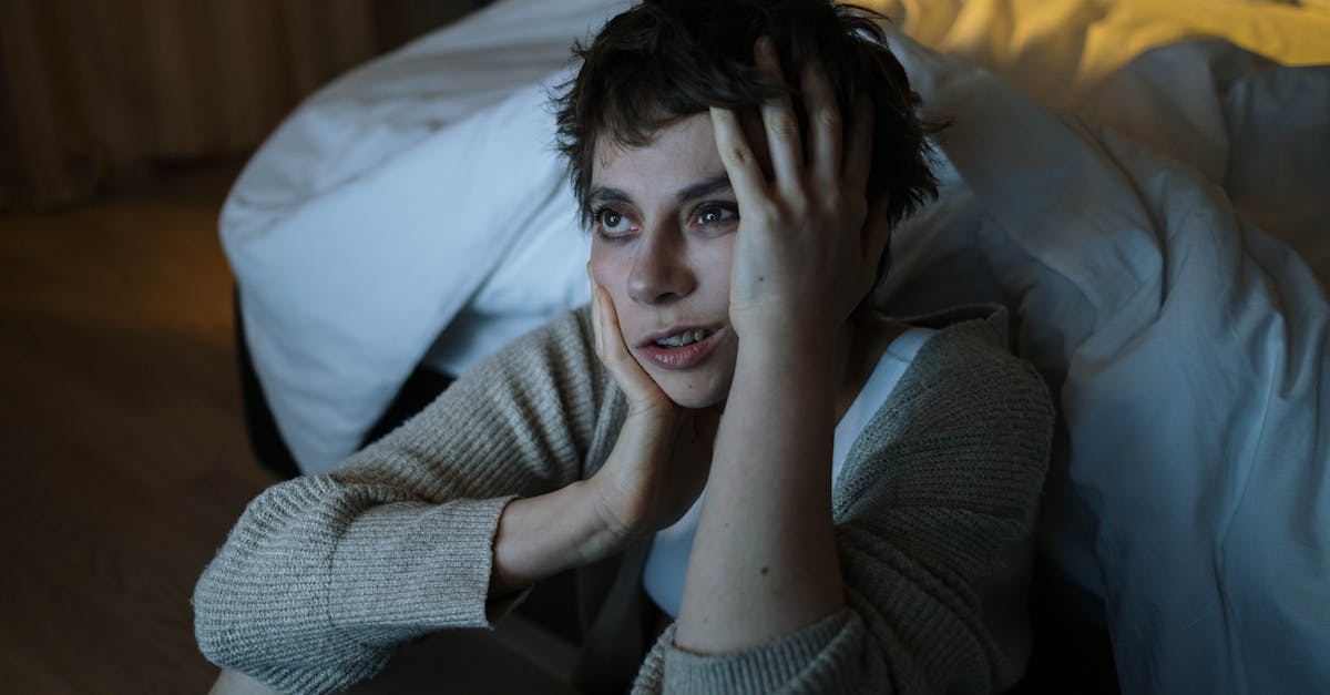 A woman sitting on the floor by a bed with a worried expression, touching her face.