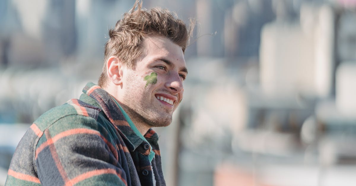 Side view of happy man with painted shamrock on cheek and glass of beer standing near railing on street against cityscape during Saint Patricks day