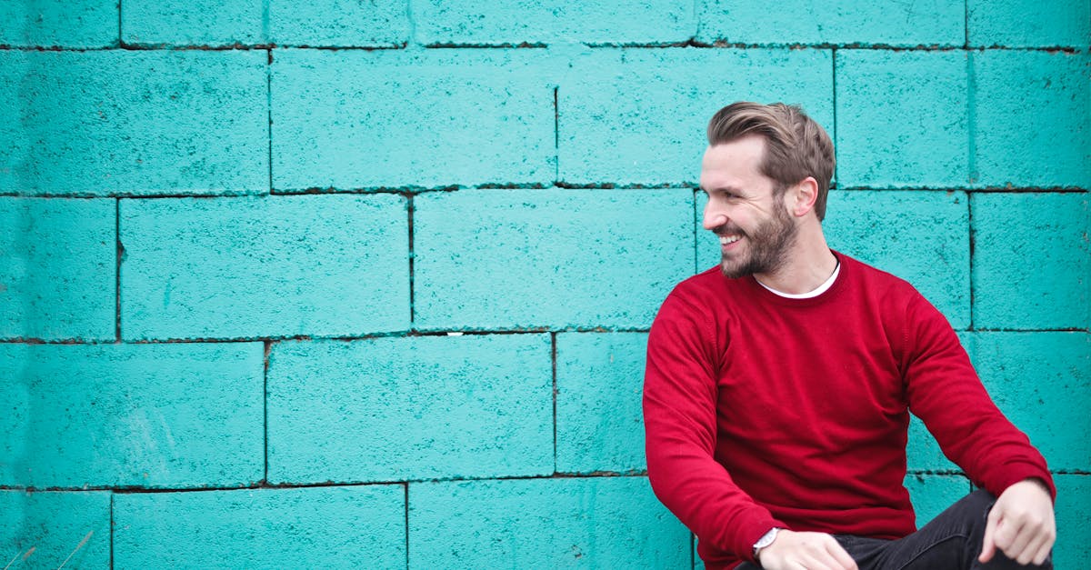 A cheerful man in a red sweater sits against a turquoise wall, enjoying a moment of happiness and laughter.