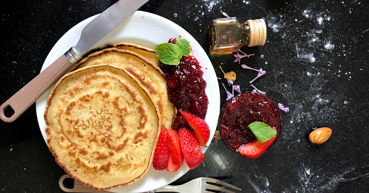 Top view of pancakes with strawberry jam, fresh strawberries, almonds, and honey on a dark kitchen counter.