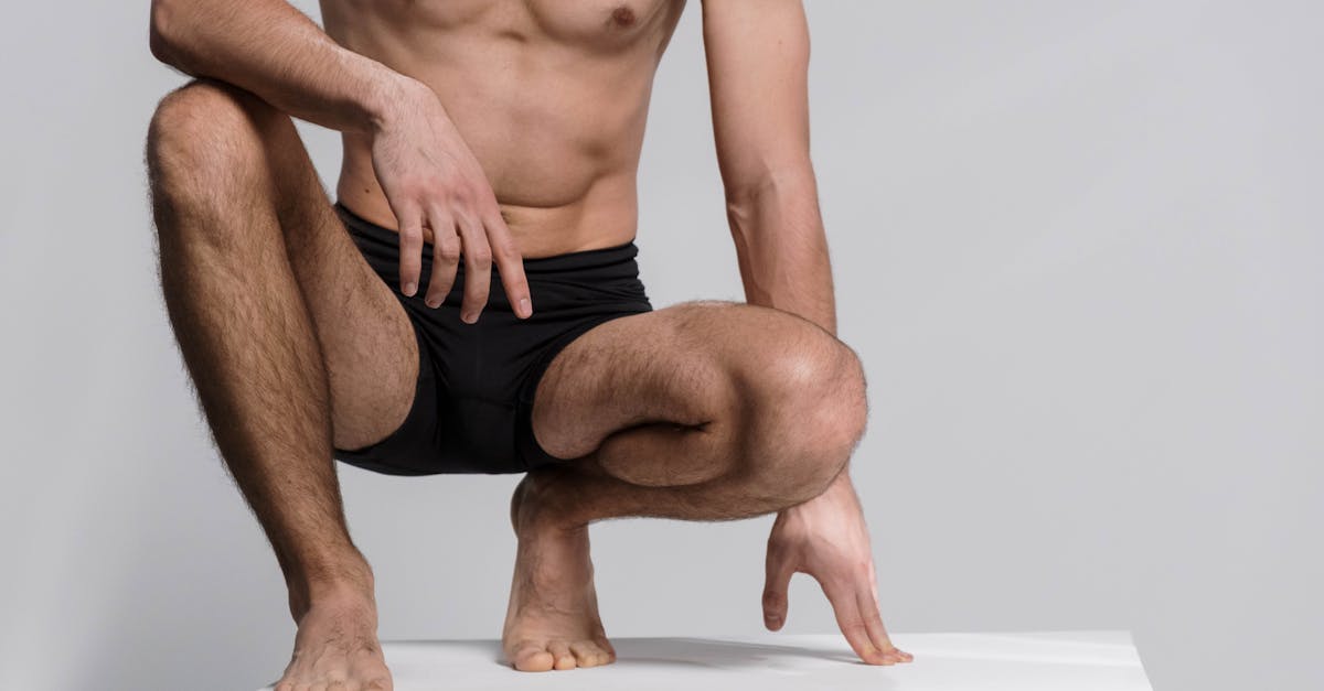 Close-up of a shirtless man squatting, wearing black underwear on a white platform.