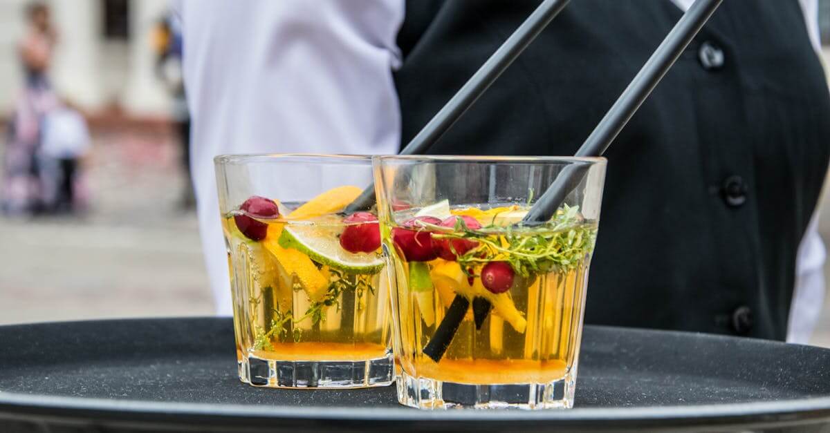 Delicious orange cocktails with cranberries and herbs on a tray, served by waiter at an outdoor event.